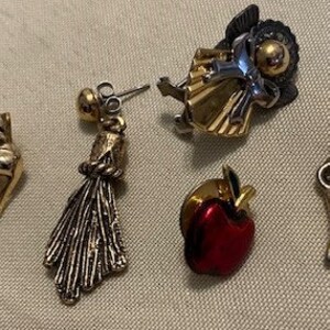 May include: Five gold-toned pins with different designs. One pin features a red apple, another a gold angel, another a gold and silver peacock, another a gold mask, and the last a gold key.