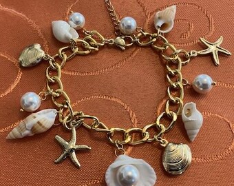 May include: Gold-toned chain bracelet adorned with seashell charms, faux pearls, and starfish pendants. The bracelet features a lobster clasp closure and is set against an orange background. The charms include various shell shapes and sizes.
