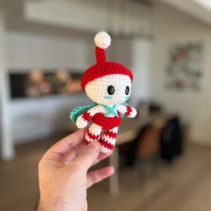 May include: A handmade crochet doll with a red hat and striped trousers. The doll has a white face with black eyes and a small blue cape. The doll is held in a person's hand.