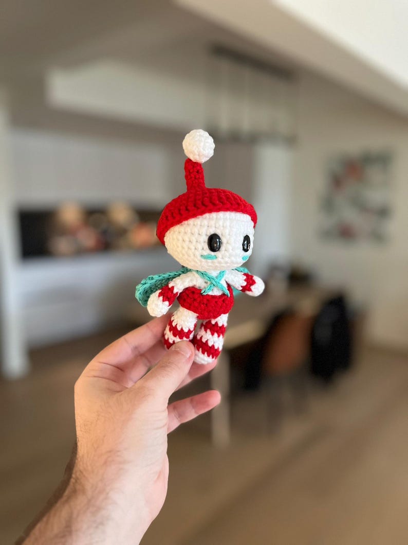May include: A handmade crochet doll with a red hat and striped trousers. The doll has a white face with black eyes and a small blue cape. The doll is held in a person's hand.