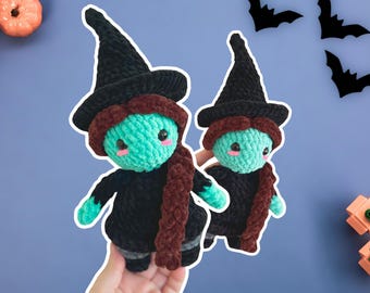 May include: Two crochet witch dolls with teal skin, black outfits, and black witch hats. They have brown hair and scarves. The dolls are held in a hand against a blue background with Halloween decorations.