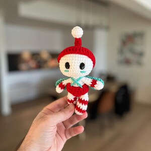 May include: A handmade crochet doll with a red hat and body, white face, and striped legs. The doll has a small white pom-pom on top of the hat and turquoise accents. The doll is a whimsical, handcrafted item.