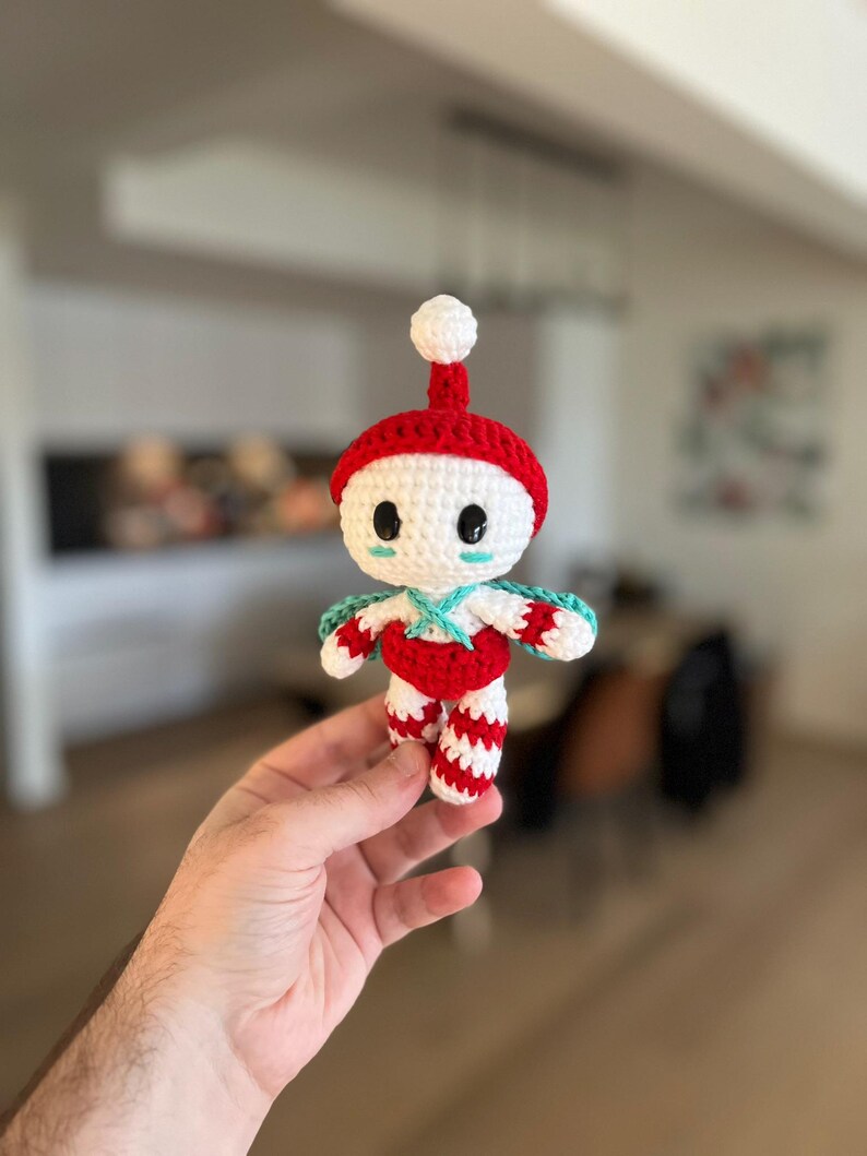May include: A handmade crochet doll with a red hat and body, white face, and striped legs. The doll has a small white pom-pom on top of the hat and turquoise accents. The doll is a whimsical, handcrafted item.