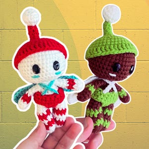 May include: Two handmade crochet dolls. One doll is white with a red hat and accents, the other is brown with a green hat and accents. Both dolls have black eyes and white wings. The dolls are held in front of a yellow and green background.