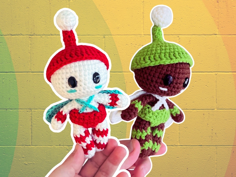 May include: Two handmade crochet dolls. One doll is white with a red hat and accents, the other is brown with a green hat and accents. Both dolls have black eyes and white wings. The dolls are held in front of a yellow and green background.