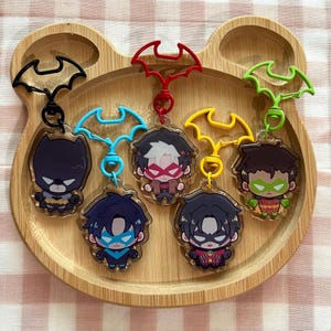 May include: Five acrylic keychains featuring chibi-style characters with bat-shaped clasps in black, blue, red, yellow, and green. The characters include a dark-suited figure, a blue-masked hero, a red-masked figure, a dark-haired hero, and a green-masked hero.