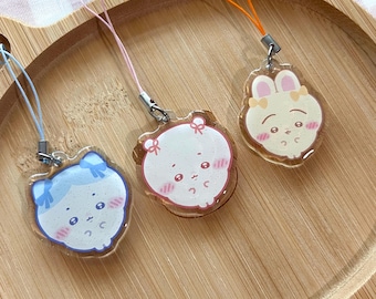 May include: Three acrylic charms featuring cartoon characters with bows, each attached to a colorful phone strap. The charms include a blue-haired character, a white character with pink accents, and a cream-colored rabbit-like character.