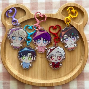 May include: Five acrylic keychains featuring chibi-style characters with large eyes and colorful hair, each attached to a heart-shaped clasp. The characters wear pajamas and headbands in various colors including purple, blue, pink, red, and yellow.
