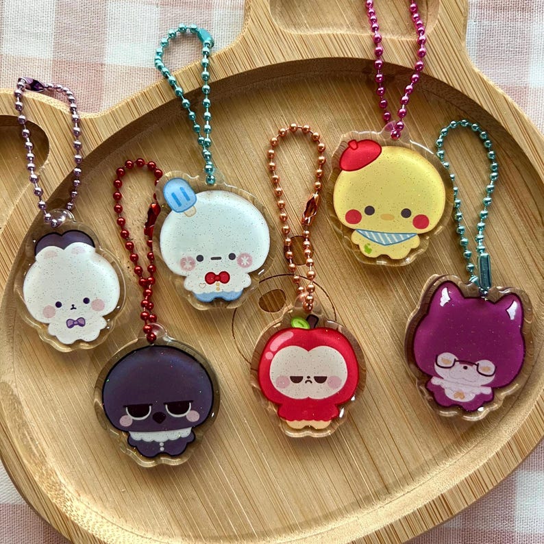 May include: Six acrylic character keychains with colorful ball chains. The charms feature cartoon figures including a white bear, a blue-capped creature, a dark purple character, a red apple-shaped figure, a yellow bird, and a purple cat.