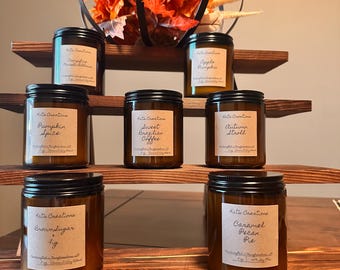 May include: A tiered wooden shelf display with a metal pumpkin decoration and eight amber glass jars with black lids. Each jar contains a candle with a label that reads "Kate Creations" and a different fall-themed scent, including "Apple Pumpkin", "Pumpkin Spice", "Sweet Brazilian Coffee", "Autumn Stroll", "Brown Sugar Fig", and "Caramel Pecan Pie".