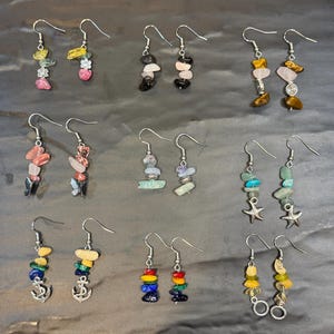 May include: Twelve pairs of dangle earrings featuring various gemstone and metal charms. The earrings are silver-toned and feature charms such as anchors, stars, and flowers.