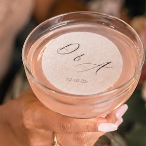 May include: A clear glass coupe filled with a pale pink cocktail, featuring a circular white paper topper printed with the black cursive text D & A and the date 10.18.25. The glass has a thin stem and a wide, rounded bowl shape.