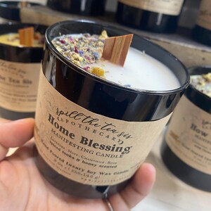 May include: A black glass candle jar with a white label that reads "Spill the Tea Sis Apothecary Home Blessing Manifesting Candle Lavender, Mint, Chamomile, Rose Spiced Tonka & Honey Scented Hand Poured Luxury Soy Wax Candle 9 oz (255.140 g)"
