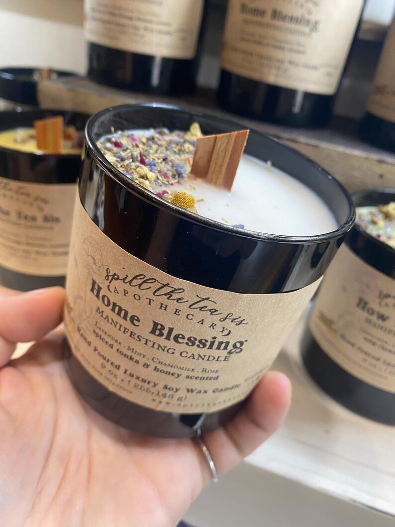 May include: A black glass candle jar with a white label that reads "Spill the Tea Sis Apothecary Home Blessing Manifesting Candle Lavender, Mint, Chamomile, Rose Spiced Tonka & Honey Scented Hand Poured Luxury Soy Wax Candle 9 oz (255.140 g)"