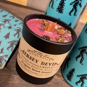 May include: A black candle jar with a red wax and a label that reads "Spill the tea sis HOME & GIFT JERSEY DEVIL Intentionally poured Cryptid Candle Devils Claw. Pine Cone. Carnelian Dragons Blood Scented 9 oz (255g)"