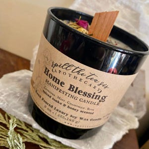 May include: A black glass candle jar with a brown label that reads "Spill the Tea Sis (Apothecary) Home Blessing Manifesting Candle Lavender, Mint, Chamomile, Rose Spiced Tonka & Honey Scented Hand Poured Luxury Soy Wax Candle 9 oz. (255.146 g)"