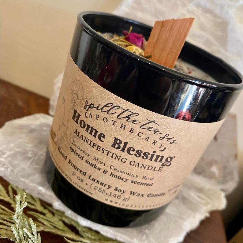 May include: A black glass candle jar with a brown label that reads "Spill the Tea Sis (Apothecary) Home Blessing Manifesting Candle Lavender, Mint, Chamomile, Rose Spiced Tonka & Honey Scented Hand Poured Luxury Soy Wax Candle 9 oz. (255.146 g)"