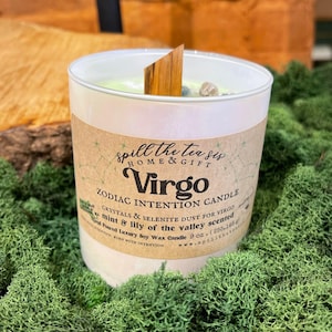 May include: A white candle with a wooden wick and a green label that says "spill the tea sis HOME & GIFT Virgo ZODIAC INTENTION CANDLE CRYSTALS & SELENITE DUST FOR VIRGO mint & lily of the valley scented Hand Poured Luxury Soy Wax Candle 9 OZ. (255.146 g) BURN WITH INTENTION www.spilltheteasis.com".