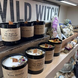 May include: A shelf display of six black glass candles with wooden wicks. The candles are labeled with the text "Spill the Tea" and various scents, including "The Magic of New Beginnings", "How Sweet It Is", and "Home Blessing".