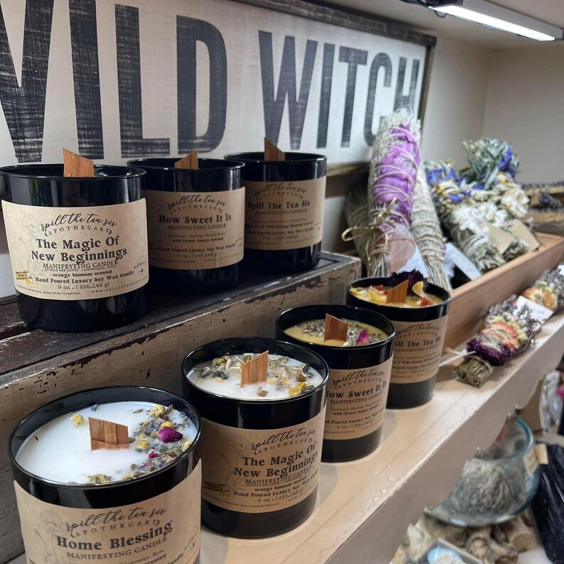 May include: A shelf display of six black glass candles with wooden wicks. The candles are labeled with the text "Spill the Tea" and various scents, including "The Magic of New Beginnings", "How Sweet It Is", and "Home Blessing".