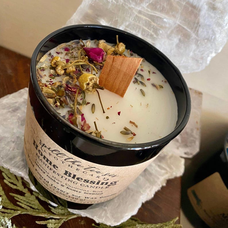 May include: A black glass candle jar with a white candle and dried flowers and herbs. The label reads "Space the Sky Apothecary Home Blessing Manifesting Candle Lavender Mint & Chamomile Hand Poured Luxury Soy Wax Candle"