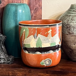 May include: An orange ceramic pot with a black rim and a decorative band featuring white houses and green trees. The pot has a spiral design on the bottom and is surrounded by other decorative items.