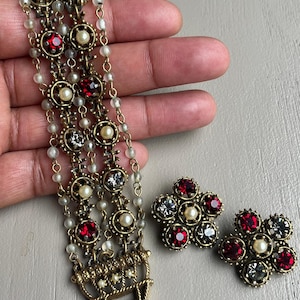 May include: Vintage jewelry set featuring a multi-strand bracelet and matching earrings. The bracelet has red, clear, and pearl-like accents. The earrings are flower-shaped with similar embellishments. The metal has a gold tone.