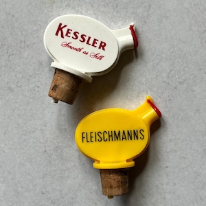 May include: Two vintage bottle stoppers. One is white with "KESSLER Smooth as Silk" in red text. The other is yellow with "FLEISCHMANN'S" in black text. Both have cork bases and red accents.