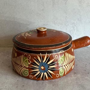May include: A brown ceramic pot with a lid and a handle. The pot is decorated with a black and white floral design and green accents.