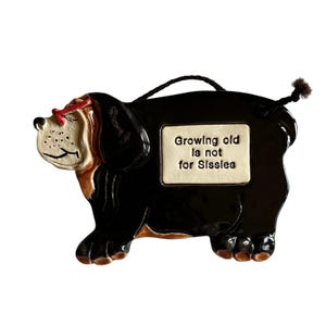 May include: A ceramic dog-shaped wall hanging with a black body, brown and tan face, and a red bow. The dog has a small rectangular sign that reads "Growing old is not for Sissies". A black string is attached for hanging.