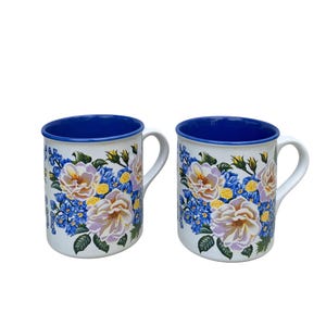 May include: Two white ceramic mugs with a blue interior and handle. The mugs are decorated with a floral pattern featuring cream-colored roses, blue forget-me-nots, and yellow accents. The rims of the mugs are also blue.