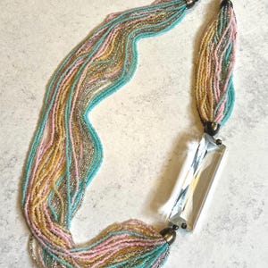 May include: A multi-strand necklace featuring numerous thin strings of small beads in shades of turquoise, pale pink, gold, and silver. The strands are gathered at both ends by metallic caps, connecting to a large, clear, rectangular glass pendant.