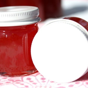 May include: Two small glass jars filled with red jelly, topped with white lids. The jars are sitting on a pink and white patterned surface.