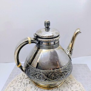 May include: A silver-toned metal teapot featuring a rounded body with an intricate embossed floral pattern around the base. It has a curved handle with light-colored accents, a matching lid with a small knob, and a slender, slightly curved spout.