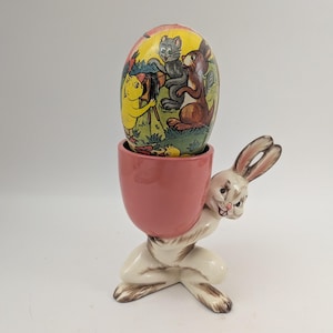 May include: A vintage Easter egg in a pink egg cup held by a white and brown bunny figurine. The egg is decorated with a colorful illustration of cartoon animals. A charming Easter decoration.