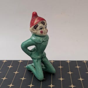 May include: A ceramic figurine of a kneeling elf with a red hat, green outfit, and rosy cheeks. The elf has a whimsical expression and is posed with hands on knees. The figurine is on a black surface with a gold star pattern.