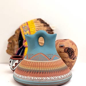 May include: A turquoise and terracotta ceramic vessel with a central handle and a wide base. The piece features etched mountain motifs, vertical line patterns, and horizontal bands in white and blue. It is displayed alongside two smaller patterned pots.