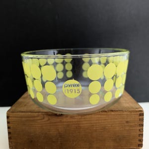 May include: A clear glass Pyrex bowl with a pattern of yellow polka dots. The bowl has a rounded shape and the text "pyrex 1915" is printed on the front. The bowl is sitting on a wooden box.