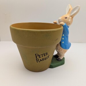May include: A tan ceramic flower pot featuring the text Peter Rabbit printed in black. A three-dimensional figure of a rabbit wearing a blue jacket is attached to the side of the pot, with its paws resting on the rim and standing on a green base.
