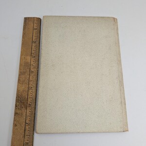 Pu&ograve; includere: Un libro d'epoca con una copertina strutturata color crema &egrave; posizionato accanto a un righello di legno. Il righello, contrassegnato in pollici, &egrave; posizionato verticalmente lungo il dorso del libro. La copertina del libro mostra segni di usura.