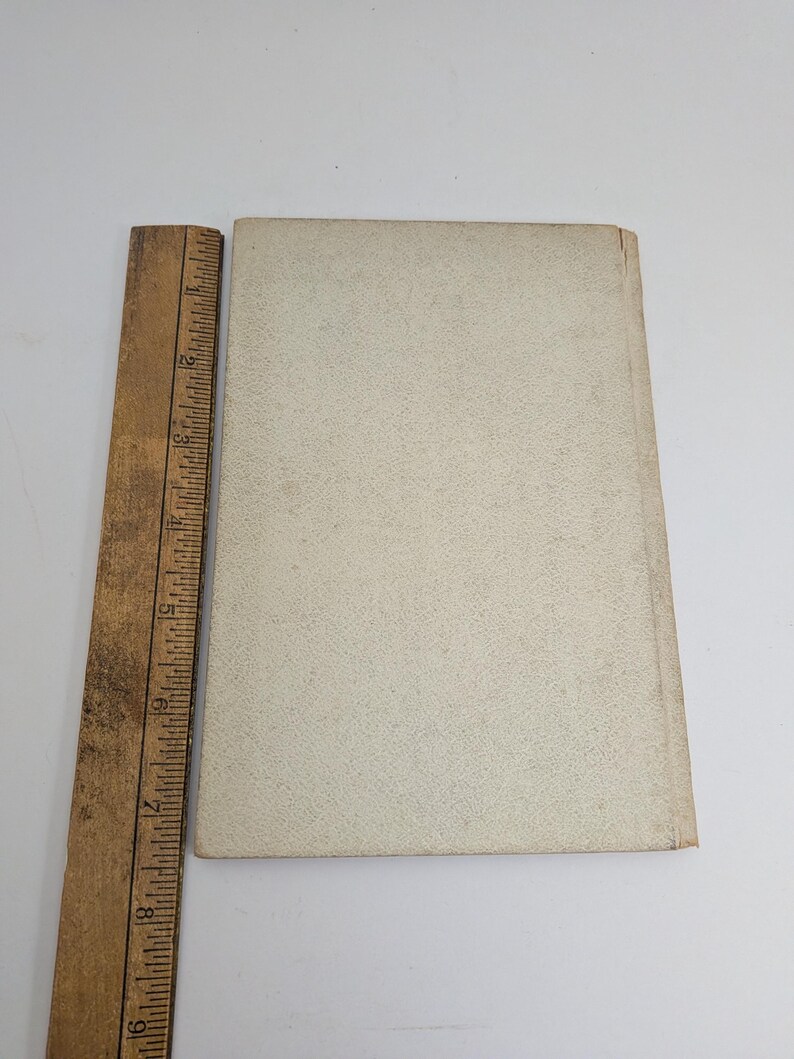 Pu&ograve; includere: Un libro d'epoca con una copertina strutturata color crema &egrave; posizionato accanto a un righello di legno. Il righello, contrassegnato in pollici, &egrave; posizionato verticalmente lungo il dorso del libro. La copertina del libro mostra segni di usura.