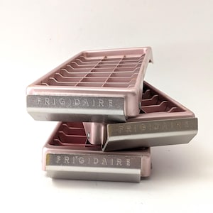 May include: Three vintage Frigidaire ice cube trays stacked on top of each other. The trays are a dusty rose color with a grid pattern and a silver metal trim. The word "FRIGIDAIRE" is printed on the front of each tray.