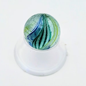 May include: A glass marble with a swirl design in shades of blue, green, and yellow. The marble is resting on a clear, circular stand. The marble's design features a mix of colors and patterns.