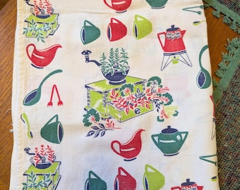 May include: A white kitchen towel with a repeating pattern of red, green, and blue kitchenware illustrations. The design includes teapots, cups, gravy boats, and a coffee grinder. The towel is folded and lies on a wooden surface.
