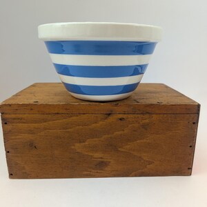 May include: A ceramic mixing bowl with a white rim and blue and white horizontal stripes. The bowl is sitting on a wooden box. The bowl is likely used for baking or cooking.