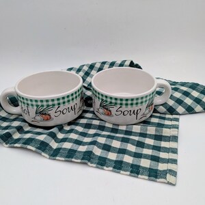 May include: Two white soup bowls with handles, featuring a green checkered pattern around the rim. The bowls are decorated with illustrations of vegetables and the words "Hot Soup". They rest on a green and white checkered cloth.