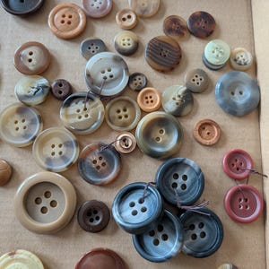 May include: A collection of various sized buttons in shades of brown, tan, and gray. The buttons have two or four holes and are made of different materials. Some buttons have a marbled appearance, while others are solid in color.