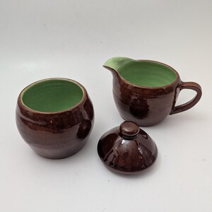 May include: A brown ceramic creamer and sugar bowl set with a matching lid. The interior of the creamer and sugar bowl are green. The set is glazed with a glossy finish.
