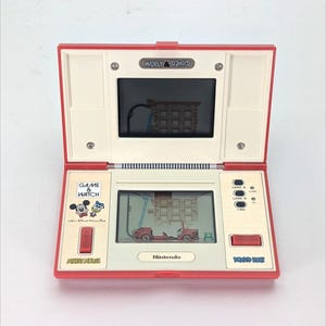 May include: A red and white Nintendo Game & Watch handheld game console featuring Mickey Mouse and Donald Duck. The game is called Mickey & Donald. The screen shows a red car driving up a ladder.