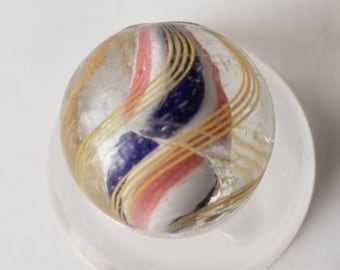 May include: A clear glass marble with internal stripes of red, white, and blue. The marble is encircled by golden-yellow swirls. The marble is resting on a clear, circular base.